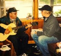 two men sitting on a couch playing guitar